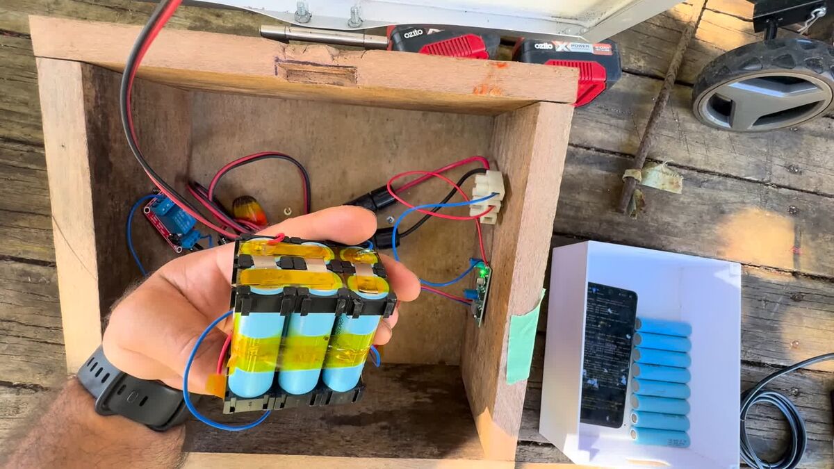 A hand holding the 3S2P pack of six 18650 cells in its holder, in front of the open plywood box with the charger and relay boards wired inside.